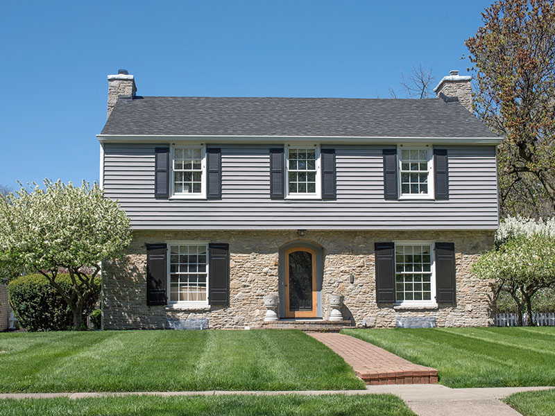 Front view of a two-story residential home with light blue vinyl siding on the top half and rocky brown brick siding on the bottom half