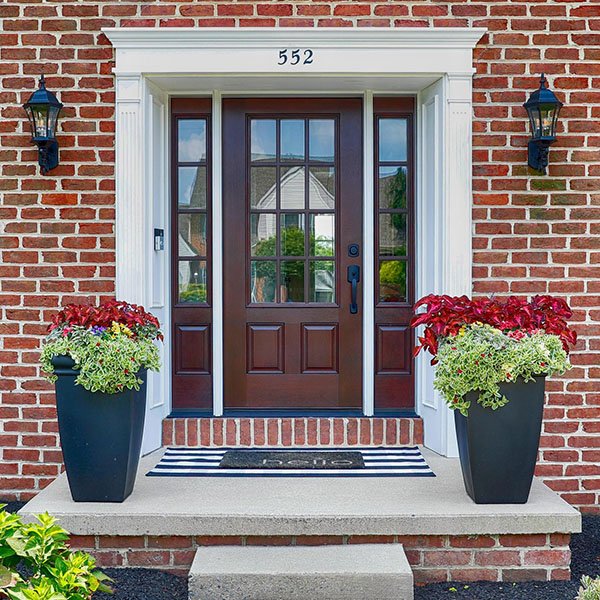 Exterior of a home and close-up of a front door made of a dark brown wood surrounded by red brick and two flower pots on either side